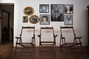 Numa sala, de piso de cimento queimado vermelho, três espreguiçadeiras de madeira, com braços, e sem a lona do assento. Acima, na parede branca, fotografias em preto e branco. Duas, em molduras ovais douradas: uma é fotopintura de um casal, e a outra, é foto de um casal, ela de noiva e ele de paletó. Abaixo, uma foto quadrada, do rosto de uma mulher de perfil em moldura dourada com quinas decoradas em relevo. Ao lado, cinco fotos grandes. A do centro é a de uma família com quatro crianças, os pais e o avô. Estão de pé. A mãe com a caçula nos braços, ambas usam vestidos. Outra menina, de uns sete anos, usa vestido com detalhes de marinheiro. O menino, de uns cinco anos, usa calça escura e camisa de malha listrada. A mais velha, de uns 11anos, está ao lado do pai, que é alto. Ele está com a mão direita no ombro dela e a esquerda no ombro da mãe. Junto ao pai, o avô. Ele é calvo, usa de camisa de botão, e está com os braços para trás. Ladeando essa foto, tem a foto individual dos filhos. À esquerda, a segunda filha; abaixo, o menino; à direita, a mais velha; e, abaixo, a caçula.