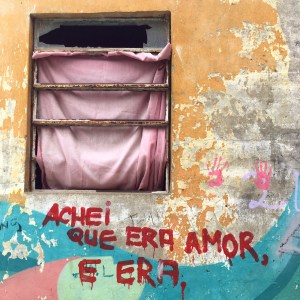 Em parte de uma fachada, um basculante, uma parede com camadas de tinta descascadas e, abaixo, pintado em vermelho: “Achei que era amor, e era.”. O basculante é enferrujado e sem vidro. Está com um lençol rosa desbotado preso por dentro na segunda abertura de cima, e passa através da penúltima abertura, com a ponta voltada para dento do cômodo. A parede tem camadas de tinta, amarelo escuro mais acima, e, por baixo, amarelo claro. Mais à direita, o reboco está à mostra; e sobre ele tem carimbos cor de rosa de duas palmas de mãos. Abaixo do basculante, a frase, que tem tinta escorrida em algumas letras e no ponto final, está sobre camadas de tintas azul, rosa e verde claro.