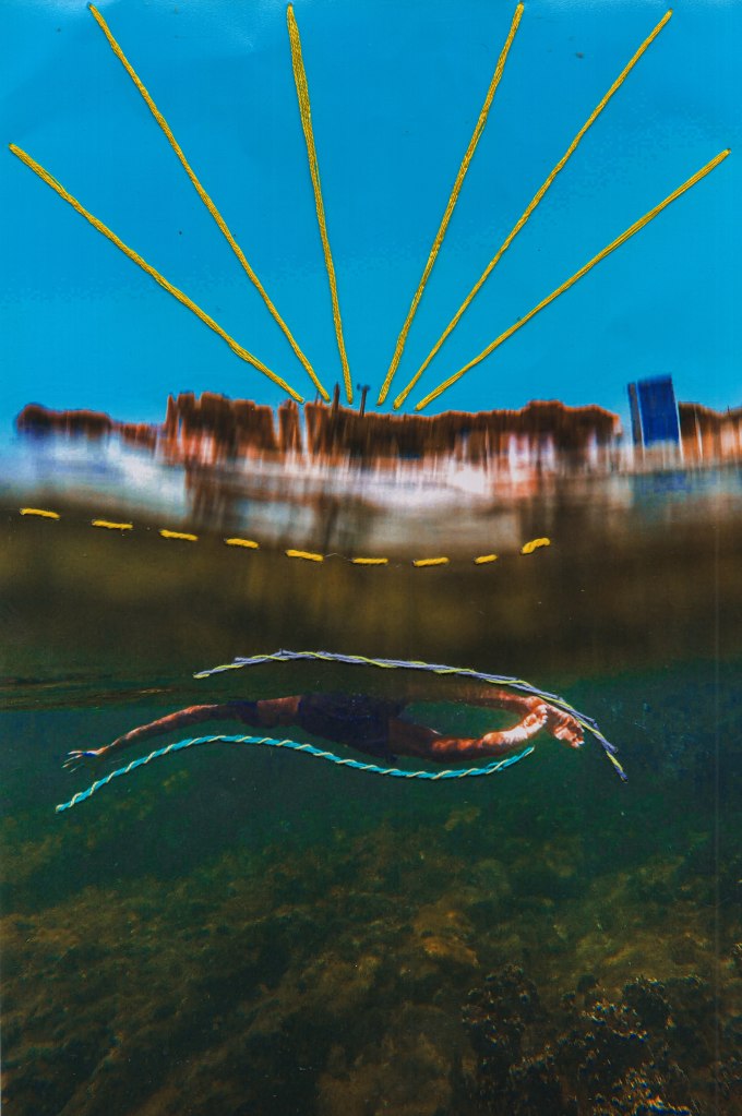 Fotografia vertical de uma paisagem com interferências digitais, como borrado, e manuais, como pespontos e alinhavos. Em primeiro plano, uma pessoa nada para a esquerda em águas esverdeadas sobre corais verde-escuros. Os pés estão próximos à superfície, ligeiramente curvos e iluminados. O corpo, visto parcialmente, é alongado.O braço esquerdo está esticado para o lado com a mão aberta. Contornando o corpo, acima, um alinhavo com linha espessa roxa, espiralada de amarelo; e, abaixo, com linha azul, também espiralada de amarelo. Acima, um pesponto amarelo parece flutuar nas águas um pouco borradas que refletem a luz da margem de uma cidade manchada com topo marrom e base intensamente iluminada. À direita, um edifício azulado. Sobre o céu azul, seis raios de linhas amarelas despontam do centro da cidade até a margem superior.
