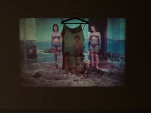 Projeção de sobreposições de fotografias em cores esmaecidas e translucidas. Duas mulheres jovens, brancas e magras, estão de pé. Usam biquini com top de faixa franzido entre os seios e com alças amarradas no pescoço. A da esquerda, na cor azul; e a da direita, em vermelho. Entre elas, no centro, dois vestidos de alças finas pendurados em cabides, a projeção de três crianças: uma em pé, de sunga vermelha; outra, sentada, olhando para a terceira, um bebê no colo de um homem, ambos parcialmente encoberto. Ao fundo, o mar com arrecifes, uma projeção que se sobrepõe ao corpo das mulheres, com as rochas refletidas nas coxas delas. Nas laterais, à esquerda, um fusquinha vermelho visto parcialmente; e, à direita, uma porta aberta. No chão, um amontoado de areia, algumas pedras e capim ralo. Uma borda preta evoca o ambiente escuro da projeção.