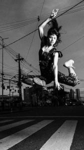 Foto em preto e branco de uma mulher jovem em pleno salto sobre uma faixa de pedestres. Ao fundo, alguns postes de eletricidade, ônibus enfileirados e prédios. Ela está com as pernas abertas e dobradas em “L” na horizontal, uma para frete e outra para trás. O braço direito esticado para cima e o esquerdo para baixo. Os cabelos longos estão levantados e as pontas tocam a mão do alto. Ela tem feições delicadas e olha para baixo com a cabeça levemente inclinada para o lado. Usa macacão estampado colado e botas.