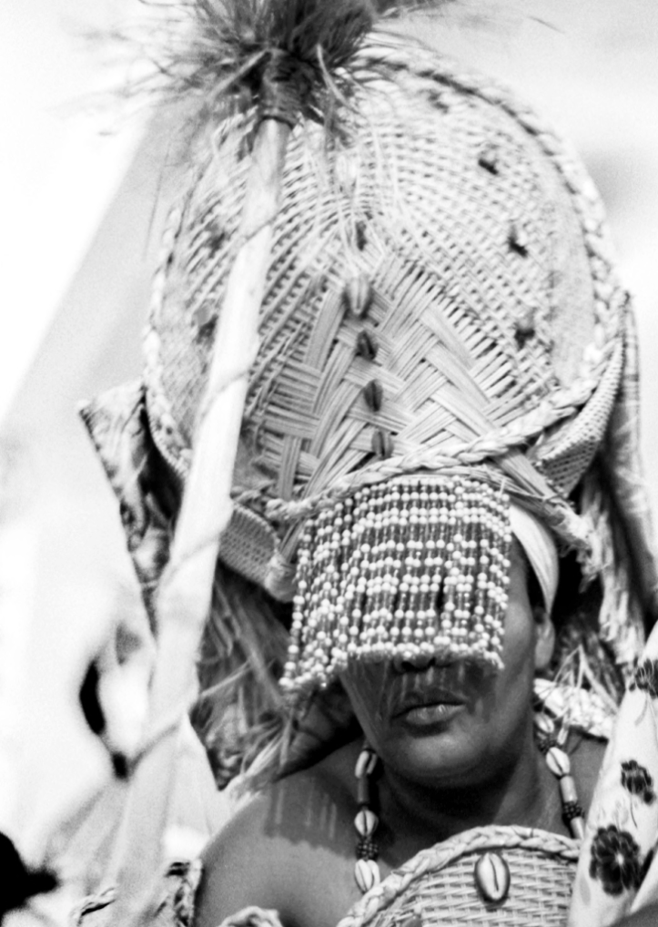 Imagem em preto e branco de uma mulher preta de lábios fartos. Ela tem um adorno em sua cabeça, como uma coroa grande de palha, que cobre seus olhos com uma franja de miçangas, e utiliza uma vestimenta feita de conchas e palha. Ela representa Oxum.